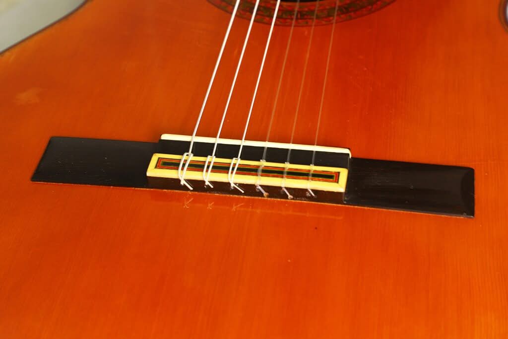 Nahaufnahme des Stegs einer „Chet Atkins Klassische Gitarre“ von Juan Estruch aus dem Jahr 1971, auf der sechs Saiten zu sehen sind, die am glänzenden orange-braunen Korpus befestigt sind, sowie ein Sattel aus hellem Holz und dekorative Einlagen. Nahaufnahme des Stegs einer „Chet Atkins Klassische Gitarre“ von Juan Estruch aus dem Jahr 1971, auf der sechs Saiten zu sehen sind, die am glänzenden orange-braunen Korpus befestigt sind, sowie ein Sattel aus hellem Holz und dekorative Einlagen.