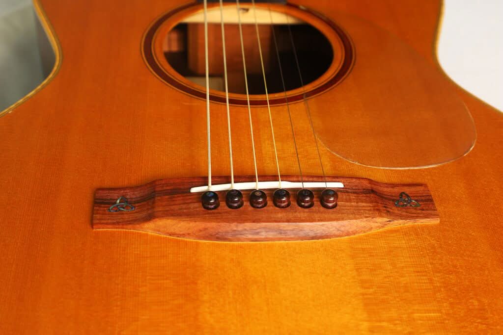 Nahaufnahme des Holzstegs der Lakewood M-54 Akustikgitarre aus dem Jahr 1998 mit sechs Saiten, die an dunklen Stiften befestigt sind, einem Teil des Schalllochs und einer glänzenden Holzoberfläche. Nahaufnahme des Holzstegs der Lakewood M-54 Akustikgitarre aus dem Jahr 1998 mit sechs Saiten, die an dunklen Stiften befestigt sind, einem Teil des Schalllochs und einer glänzenden Holzoberfläche.