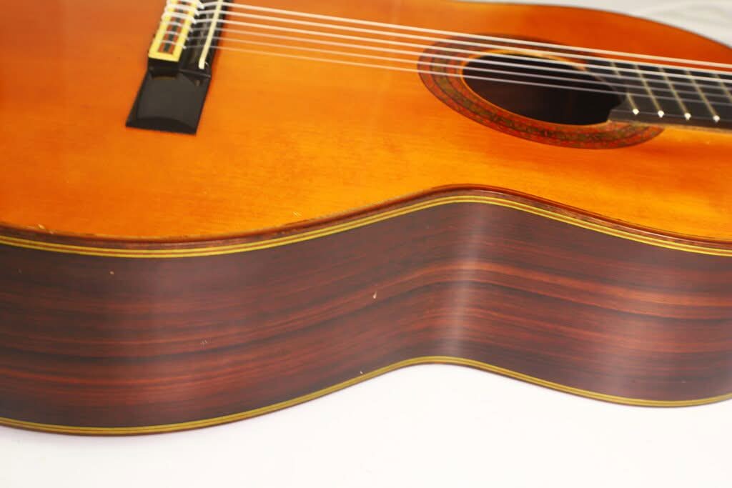 Nahaufnahme des Korpus einer „Chet Atkins Klassische Gitarre“ von Juan Estruch aus dem Jahr 1971, bei der die Holzmaserung, die Saiten und ein Teil des Schalllochs vor einem einfarbigen Hintergrund hervorgehoben werden. Nahaufnahme des Korpus einer „Chet Atkins Klassische Gitarre“ von Juan Estruch aus dem Jahr 1971, bei der die Holzmaserung, die Saiten und ein Teil des Schalllochs vor einem einfarbigen Hintergrund hervorgehoben werden.