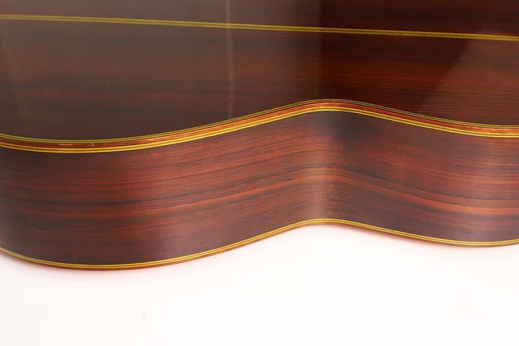 Nahaufnahme der Seite einer „Chet Atkins Klassische Gitarre“ von Juan Estruch aus dem Jahr 1971 mit einer glatten, glänzenden Lackierung, natürlicher Holzmaserung sowie gelben und schwarzen Zierkanten. Nahaufnahme der Seite einer „Chet Atkins Klassische Gitarre“ von Juan Estruch aus dem Jahr 1971 mit einer glatten, glänzenden Lackierung, natürlicher Holzmaserung sowie gelben und schwarzen Zierkanten.