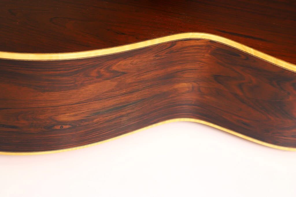 Nahaufnahme der Seite einer Lakewood M-54 Akustikgitarre aus dem Jahr 1998, die vor einem hellen Hintergrund ihre satte braune Holzmaserung und die hellen Zierleisten am Rand zur Geltung bringt. Nahaufnahme der Seite einer Lakewood M-54 Akustikgitarre aus dem Jahr 1998, die vor einem hellen Hintergrund ihre satte braune Holzmaserung und die hellen Zierleisten am Rand zur Geltung bringt.