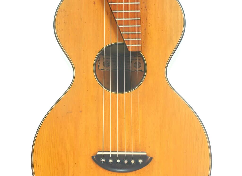 Nahaufnahme der Georg-Haid-Terz-Gitarre von 1936 mit hellbrauner Holzoberfläche, sechs Saiten und einem runden Schallloch, fotografiert vor weißem Hintergrund. Nahaufnahme der Georg-Haid-Terz-Gitarre von 1936 mit hellbrauner Holzoberfläche, sechs Saiten und einem runden Schallloch, fotografiert vor weißem Hintergrund.