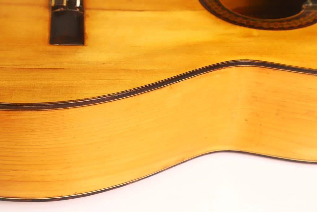 Nahaufnahme des Korpus der Akustikgitarre „Modesto Borreguero 1955“, die die glatte, helle Holzoberfläche und die dunklen Zierleisten hervorhebt und einen Teil des Schalllochs sowie den Steg zeigt. Nahaufnahme des Korpus der Akustikgitarre „Modesto Borreguero 1955“, die die glatte, helle Holzoberfläche und die dunklen Zierleisten hervorhebt und einen Teil des Schalllochs sowie den Steg zeigt.