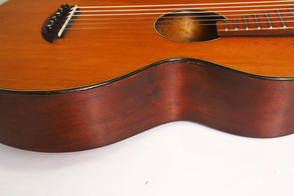 Nahaufnahme des Korpus einer Terz-Gitarre von Georg Haid aus dem Jahr 1936, die die Holzmaserung, die Saiten und einen Teil des Schalllochs vor weißem Hintergrund hervorhebt. Nahaufnahme des Korpus einer Terz-Gitarre von Georg Haid aus dem Jahr 1936, die die Holzmaserung, die Saiten und einen Teil des Schalllochs vor weißem Hintergrund hervorhebt.
