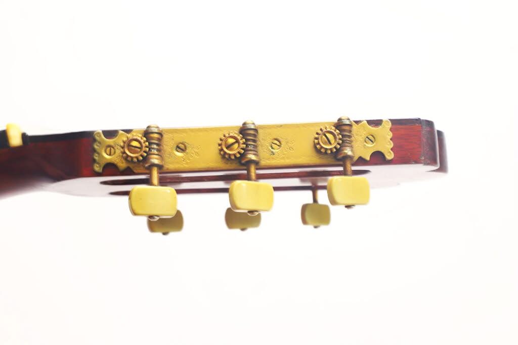 Nahaufnahme des Kopfes einer „Francisco Simplicio 1931 Torres Klassische Gitarre“ mit gelben Stimmwirbeln und verzierten Messingbeschlägen vor weißem Hintergrund. Nahaufnahme des Kopfes einer „Francisco Simplicio 1931 Torres Klassische Gitarre“ mit gelben Stimmwirbeln und verzierten Messingbeschlägen vor weißem Hintergrund.