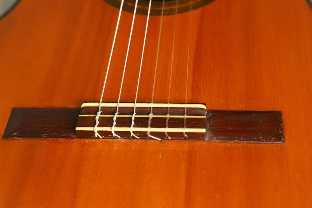 Nahaufnahme des Holzstegs und der Saiten der „Francisco Simplicio 1931 Torres Klassische Gitarre“ auf ihrem hellbraun polierten Korpus, wobei besonders deutlich zu sehen ist, wo die Saiten am Steg befestigt und verankert sind. Nahaufnahme des Holzstegs und der Saiten der „Francisco Simplicio 1931 Torres Klassische Gitarre“ auf ihrem hellbraun polierten Korpus, wobei besonders deutlich zu sehen ist, wo die Saiten am Steg befestigt und verankert sind.