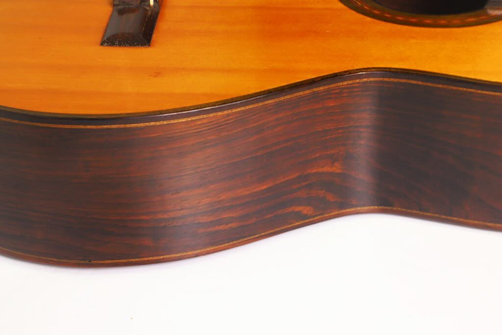 Nahaufnahme der Seite der „Francisco Simplicio 1931 Torres Klassische Gitarre“, die die glatte, dunkle Holzmaserung und die warme, glänzende Lackierung vor einem hellen Hintergrund hervorhebt. Nahaufnahme der Seite der „Francisco Simplicio 1931 Torres Klassische Gitarre“, die die glatte, dunkle Holzmaserung und die warme, glänzende Lackierung vor einem hellen Hintergrund hervorhebt.