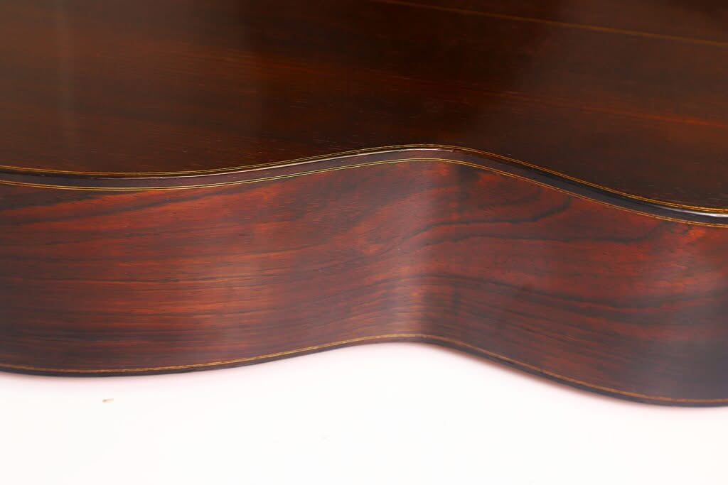 Nahaufnahme der Seite einer „Torres Klassische Gitarre“ von Francisco Simplicio aus dem Jahr 1931, die die satte, dunkle Holzmaserung und die eleganten Rundungen vor einem hellen Hintergrund hervorhebt. Nahaufnahme der Seite einer „Torres Klassische Gitarre“ von Francisco Simplicio aus dem Jahr 1931, die die satte, dunkle Holzmaserung und die eleganten Rundungen vor einem hellen Hintergrund hervorhebt.