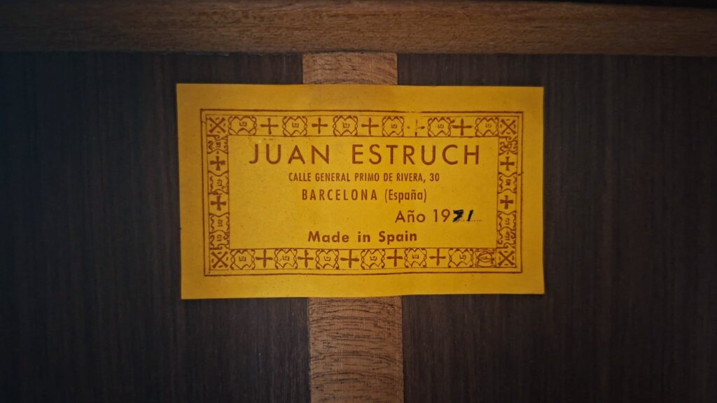 Auf einem gelben Etikett auf dunklem Holz steht: „Juan Estruch 1971 Chet Atkins Klassische Gitarre, Calle General Primo de Rivera 30, Barcelona (Spanien), Jahr 1971, Made in Spain“, umrahmt von einer Zierleiste. Auf einem gelben Etikett auf dunklem Holz steht: „Juan Estruch 1971 Chet Atkins Klassische Gitarre, Calle General Primo de Rivera 30, Barcelona (Spanien), Jahr 1971, Made in Spain“, umrahmt von einer Zierleiste.