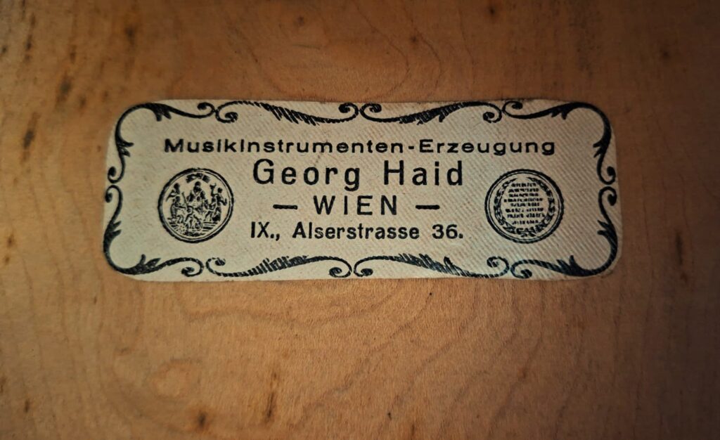 Auf einem Vintage-Etikett der Georg-Haid-Terz-Gitarre von 1936 steht „MusikInstrumenten-Erzeugung Georg Haid WIEN, IX., Alserstraße 36“, umrahmt von zwei runden Emblemen auf jeder Seite, die auf der Holzoberfläche angebracht sind. Auf einem Vintage-Etikett der Georg-Haid-Terz-Gitarre von 1936 steht „MusikInstrumenten-Erzeugung Georg Haid WIEN, IX., Alserstraße 36“, umrahmt von zwei runden Emblemen auf jeder Seite, die auf der Holzoberfläche angebracht sind.