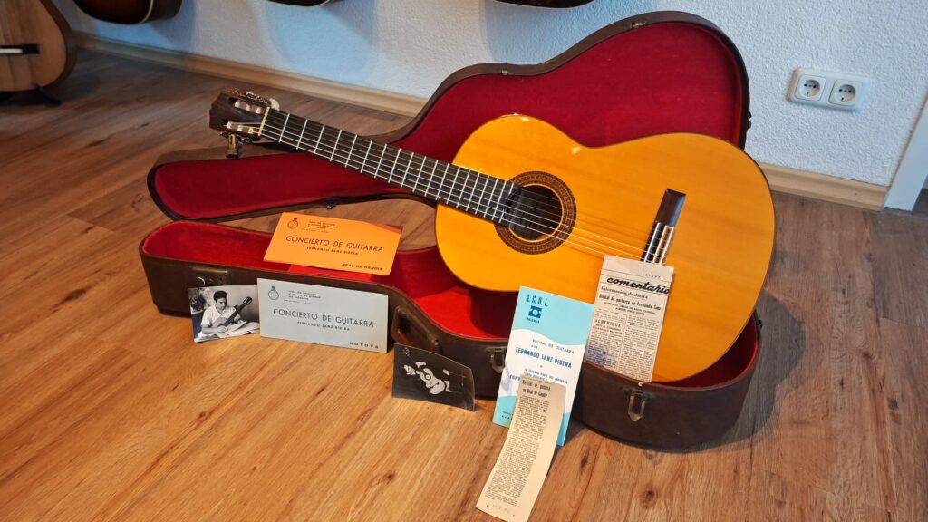 Eine Flamenco-Gitarre der Marke „Sobrinos de Santos Hernandez“ aus dem Jahr 1974 liegt in einem offenen Hartschalenkoffer auf einem Holzboden, umgeben von Konzertprogrammheften und Papieren, vor einer weißen Wand mit zwei Steckdosen. Eine Flamenco-Gitarre der Marke „Sobrinos de Santos Hernandez“ aus dem Jahr 1974 liegt in einem offenen Hartschalenkoffer auf einem Holzboden, umgeben von Konzertprogrammheften und Papieren, vor einer weißen Wand mit zwei Steckdosen.