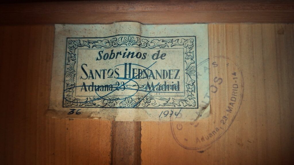Auf einem alten Etikett im Inneren der Flamenco-Gitarre „Sobrinos de Santos Hernandez 1974“ steht „Sobrinos de Santos Hernandez, Aduana 23, Madrid“; rechts sind eine Unterschrift, ein Datum und ein teilweise sichtbarer runder Stempel zu sehen. Auf einem alten Etikett im Inneren der Flamenco-Gitarre „Sobrinos de Santos Hernandez 1974“ steht „Sobrinos de Santos Hernandez, Aduana 23, Madrid“; rechts sind eine Unterschrift, ein Datum und ein teilweise sichtbarer runder Stempel zu sehen.
