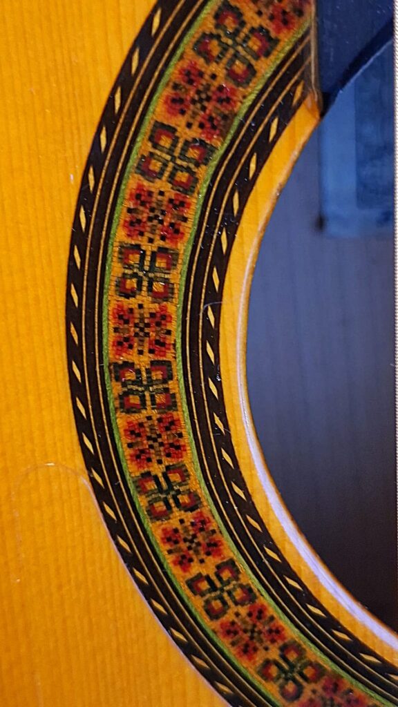 Nahaufnahme des Schalllochs der Flamenco-Gitarre „Sobrinos de Santos Hernandez 1974“, die die filigranen geometrischen Muster in Rot, Gelb, Schwarz und Grün hervorhebt, die den Rand des Holzes zieren. Nahaufnahme des Schalllochs der Flamenco-Gitarre „Sobrinos de Santos Hernandez 1974“, die die filigranen geometrischen Muster in Rot, Gelb, Schwarz und Grün hervorhebt, die den Rand des Holzes zieren.