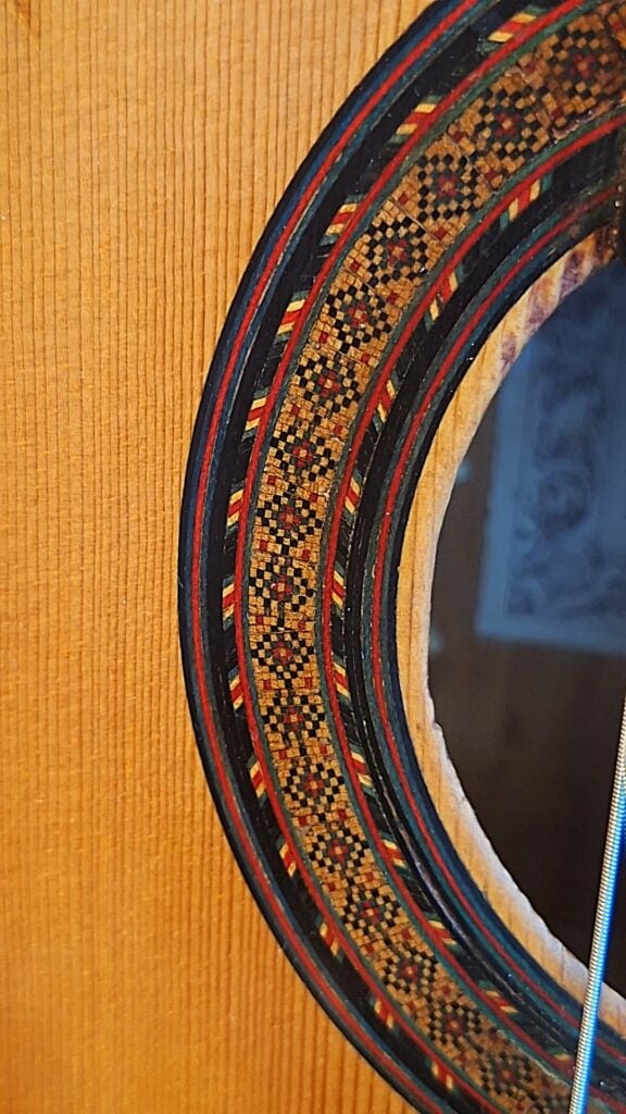 Nahaufnahme des Schalllochs der Modesto-Borreguero-Gitarre von 1955, die eine lebhafte geometrische Rosette auf heller Holzmaserung zeigt, rechts davon eine Gitarrensaite. Nahaufnahme des Schalllochs der Modesto-Borreguero-Gitarre von 1955, die eine lebhafte geometrische Rosette auf heller Holzmaserung zeigt, rechts davon eine Gitarrensaite.