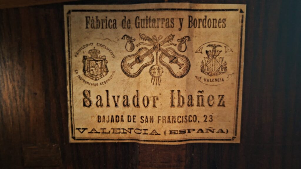 Auf einem Vintage-Etikett auf Holz für die Salvador Ibáñez 1900 Klassische Gitarre steht „Fábrica de Guitarras y Bordones Salvador Ibáñez, Bajada de San Francisco, 23, Valencia (España)“, verziert mit kunstvollen Gitarrenmotiven und Wappen. Auf einem Vintage-Etikett auf Holz für die Salvador Ibáñez 1900 Klassische Gitarre steht „Fábrica de Guitarras y Bordones Salvador Ibáñez, Bajada de San Francisco, 23, Valencia (España)“, verziert mit kunstvollen Gitarrenmotiven und Wappen.
