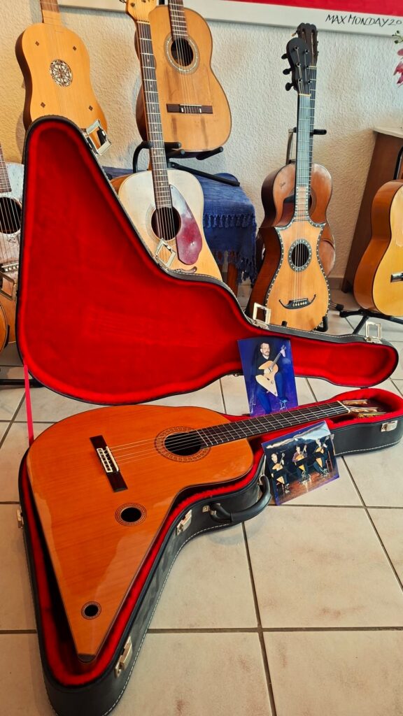 Die „Gran Klassische Gitarre“ von Raimundo und Aparicio Corujo ruht in einem offenen roten Kasten auf einem Fliesenboden, daneben liegen Fotos eines Musikers, während im Hintergrund mehrere Akustikgitarren auf Ständern ausgestellt sind. Die „Gran Klassische Gitarre“ von Raimundo und Aparicio Corujo ruht in einem offenen roten Kasten auf einem Fliesenboden, daneben liegen Fotos eines Musikers, während im Hintergrund mehrere Akustikgitarren auf Ständern ausgestellt sind.