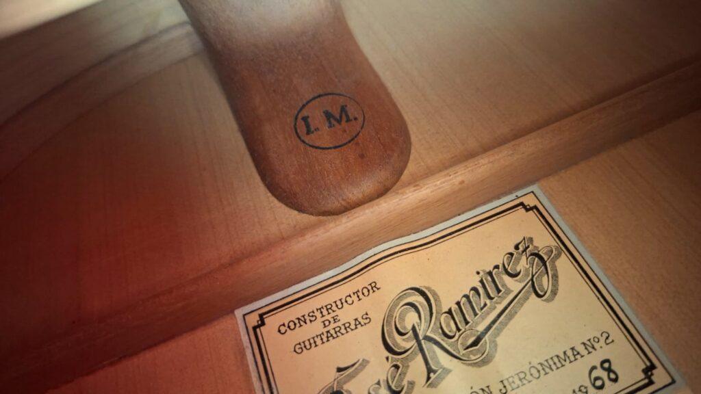 Nahaufnahme des Innenraums einer Flamenco-Gitarre vom Typ Jose Ramirez 1a, Baujahr 1968, die einen Stempel mit der Aufschrift „I.M.“ auf dem Holz sowie ein teilweise sichtbares Etikett mit der Aufschrift „Constructor de Guitarras, José Ramírez“ zeigt. Nahaufnahme des Innenraums einer Flamenco-Gitarre vom Typ Jose Ramirez 1a, Baujahr 1968, die einen Stempel mit der Aufschrift „I.M.“ auf dem Holz sowie ein teilweise sichtbares Etikett mit der Aufschrift „Constructor de Guitarras, José Ramírez“ zeigt.
