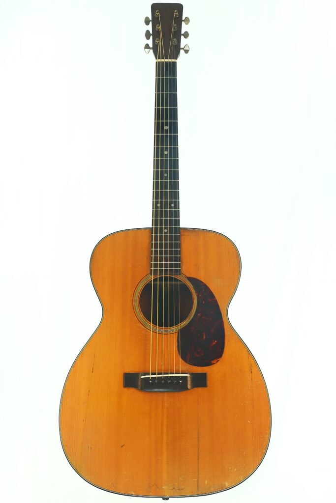 Die Akustikgitarre Martin 000-18 aus dem Jahr 1960 mit naturbelassener Holzoberfläche, dunklem Schlagbrett und sechs Saiten wird vor einem schlichten weißen Hintergrund präsentiert. Die Akustikgitarre Martin 000-18 aus dem Jahr 1960 mit naturbelassener Holzoberfläche, dunklem Schlagbrett und sechs Saiten wird vor einem schlichten weißen Hintergrund präsentiert.