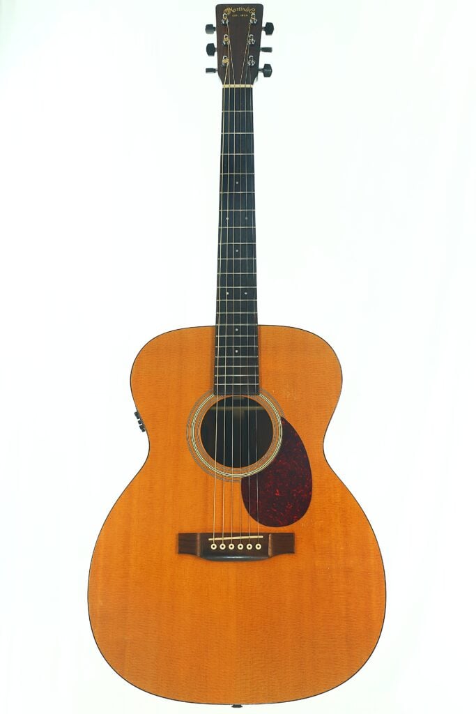Eine Gesamtansicht der MARTIN OM-21 Akustikgitarre aus dem Jahr 1995 mit naturbelassener Holzoberfläche, schwarzem Schlagbrett und sechs Saiten, fotografiert vor einem schlichten weißen Hintergrund. Eine Gesamtansicht der MARTIN OM-21 Akustikgitarre aus dem Jahr 1995 mit naturbelassener Holzoberfläche, schwarzem Schlagbrett und sechs Saiten, fotografiert vor einem schlichten weißen Hintergrund.