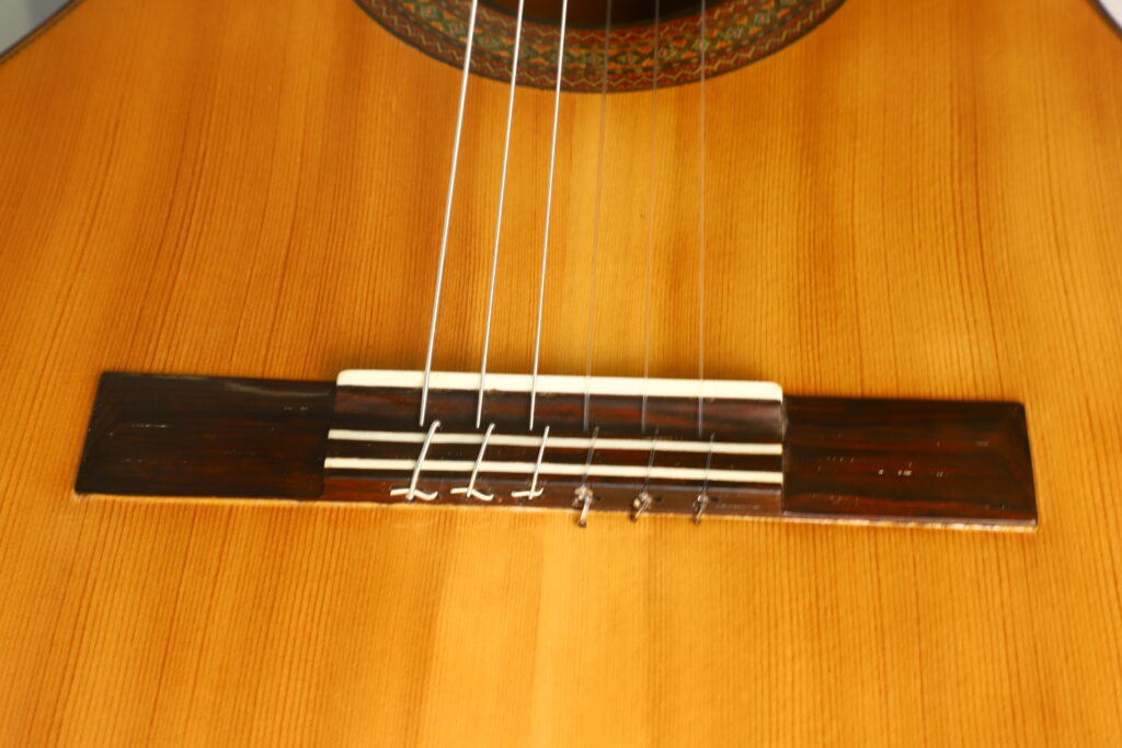 Nahaufnahme des unteren Teils einer „Francisco Klassische Gitarre“ von Manuel Adalid Sr. aus dem Jahr 1971, wobei der Steg, der Sattel, die Saiten und ein Abschnitt des naturbelassenen Holzkörpers besonders hervorgehoben sind. Nahaufnahme des unteren Teils einer „Francisco Klassische Gitarre“ von Manuel Adalid Sr. aus dem Jahr 1971, wobei der Steg, der Sattel, die Saiten und ein Abschnitt des naturbelassenen Holzkörpers besonders hervorgehoben sind.