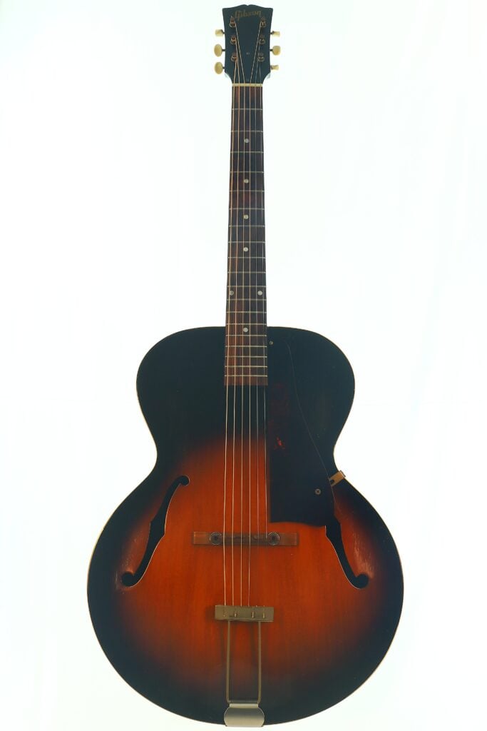 Eine Gibson L-48 Akustik Gitarre aus dem Jahr 1948 im Sunburst-Archtop-Design mit F-Löchern, einem dunklen Außenrand, der in einen helleren braunen Mittelbereich übergeht, und einem schwarzen Schlagbrett, abgebildet vor weißem Hintergrund. Eine Gibson L-48 Akustik Gitarre aus dem Jahr 1948 im Sunburst-Archtop-Design mit F-Löchern, einem dunklen Außenrand, der in einen helleren braunen Mittelbereich übergeht, und einem schwarzen Schlagbrett, abgebildet vor weißem Hintergrund.