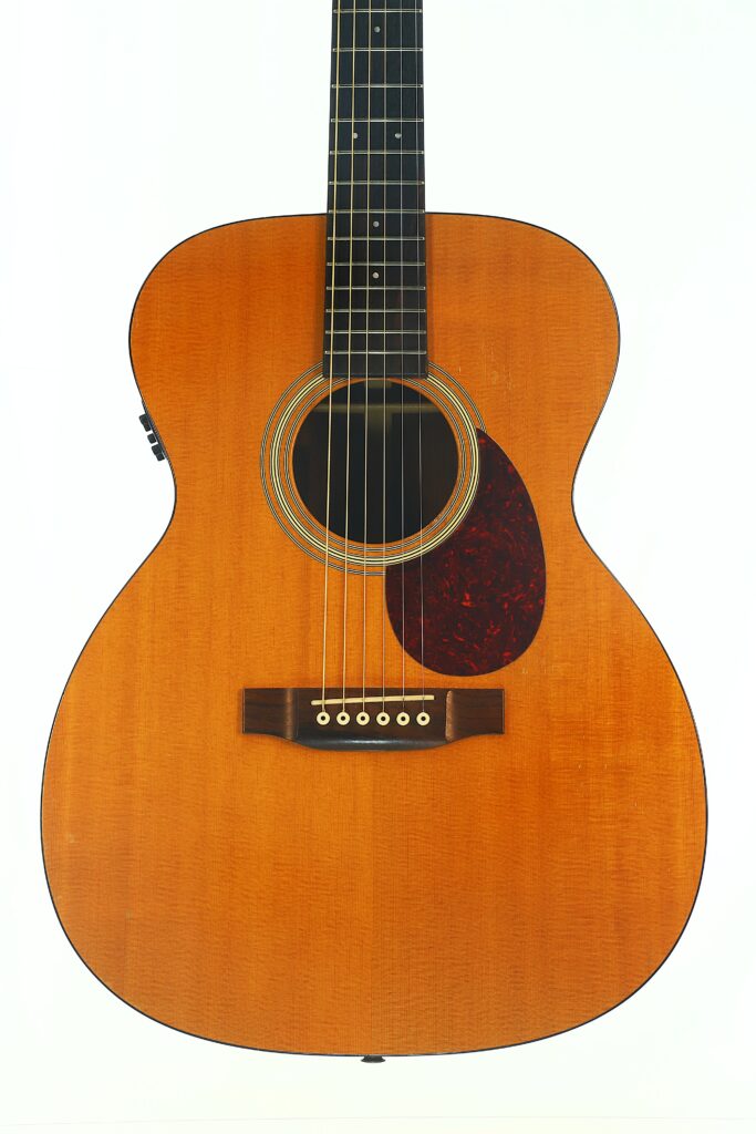 Eine Nahaufnahme der MARTIN OM-21 Akustikgitarre aus dem Jahr 1995 mit naturbelassener Holzoberfläche, auf der der Korpus, die Saiten, das Schallloch und das Schlagbrett vor einem weißen Hintergrund hervorgehoben sind. Eine Nahaufnahme der MARTIN OM-21 Akustikgitarre aus dem Jahr 1995 mit naturbelassener Holzoberfläche, auf der der Korpus, die Saiten, das Schallloch und das Schlagbrett vor einem weißen Hintergrund hervorgehoben sind.