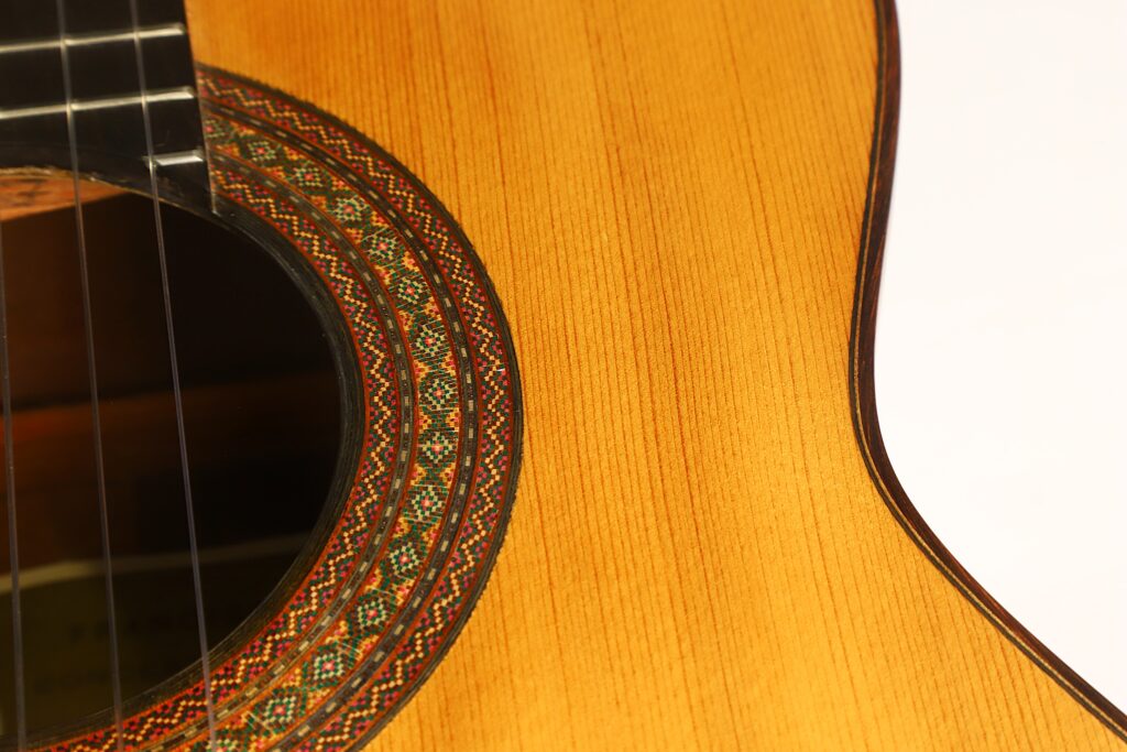 Nahaufnahme einer „Francisco Klassische Gitarre“ von Manuel Adalid Sr. aus dem Jahr 1971, die die aufwendig verzierte Rosette um das Schallloch sowie die wunderschöne Maserung des Holzkorpus hervorhebt. Nahaufnahme einer „Francisco Klassische Gitarre“ von Manuel Adalid Sr. aus dem Jahr 1971, die die aufwendig verzierte Rosette um das Schallloch sowie die wunderschöne Maserung des Holzkorpus hervorhebt.