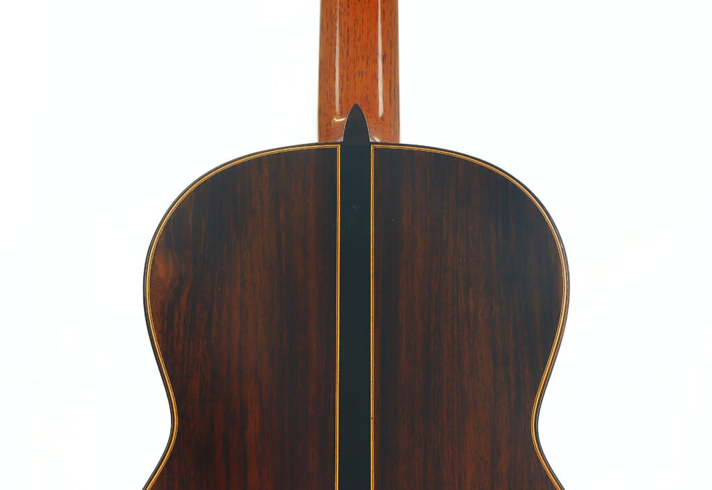 Das Bild zeigt die Rückseite einer „Friederich Klassische Gitarre“ von Stefan Nitschke aus dem Jahr 2015, die sich durch dunkles Holz mit glänzender Oberfläche und eine schmale gelbe Zierlinie in der Mitte auszeichnet. Das Bild zeigt die Rückseite einer „Friederich Klassische Gitarre“ von Stefan Nitschke aus dem Jahr 2015, die sich durch dunkles Holz mit glänzender Oberfläche und eine schmale gelbe Zierlinie in der Mitte auszeichnet.