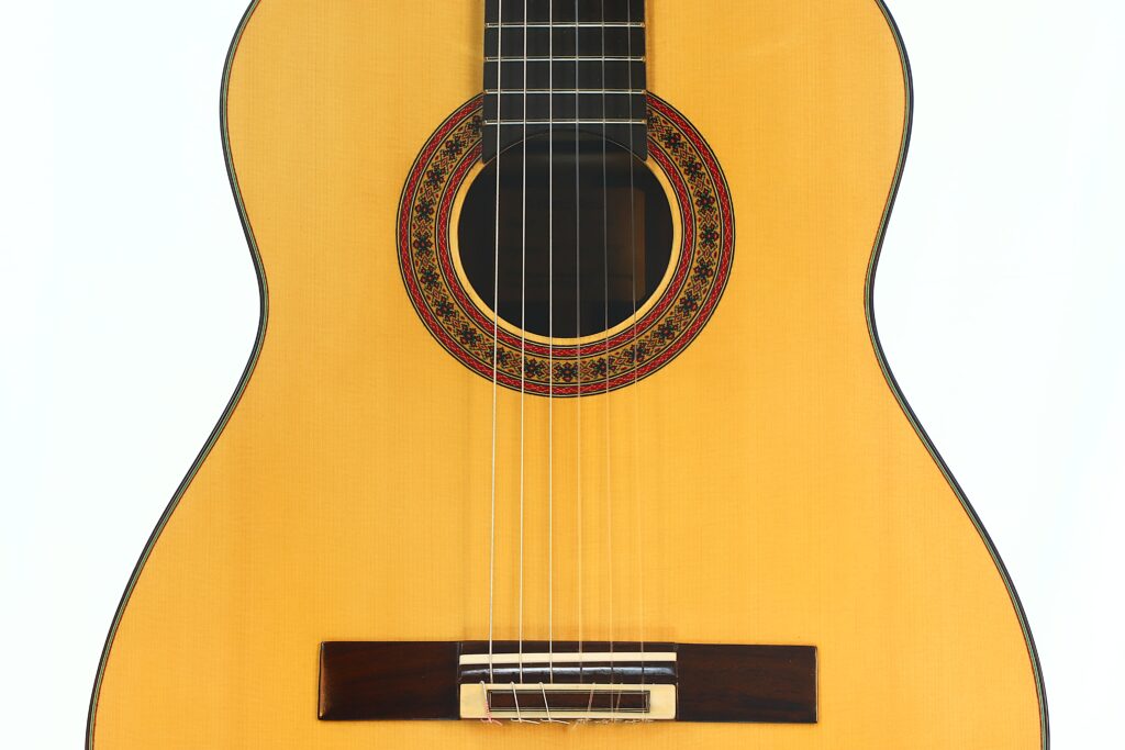 Nahaufnahme des Korpus der „Gerhard Arns 2026 Smallman Klassische Gitarre“, die das Schallloch mit der Zierrosette, die Saiten, den Steg und einen Teil des Griffbretts vor weißem Hintergrund zeigt. Nahaufnahme des Korpus der „Gerhard Arns 2026 Smallman Klassische Gitarre“, die das Schallloch mit der Zierrosette, die Saiten, den Steg und einen Teil des Griffbretts vor weißem Hintergrund zeigt.