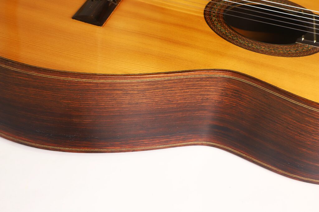 Nahaufnahme der Seite und Oberseite einer „Francisco Klassische Gitarre“ von Manuel Adalid Sr. aus dem Jahr 1971, die die Holzstruktur und -maserung, die Saiten sowie einen Teil des Schalllochs mit dekorativer Einlage hervorhebt. Der Hintergrund ist schlicht weiß. Nahaufnahme der Seite und Oberseite einer „Francisco Klassische Gitarre“ von Manuel Adalid Sr. aus dem Jahr 1971, die die Holzstruktur und -maserung, die Saiten sowie einen Teil des Schalllochs mit dekorativer Einlage hervorhebt. Der Hintergrund ist schlicht weiß.