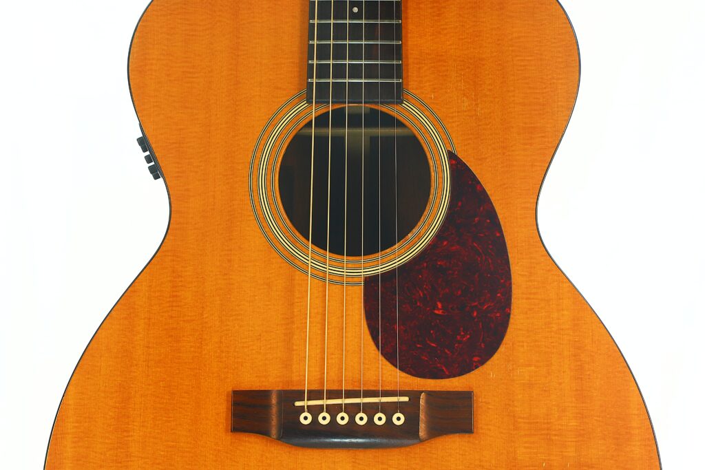 Nahaufnahme des Korpus einer MARTIN OM-21 Akustikgitarre aus dem Jahr 1995, wobei das Schallloch, die Saiten, der Steg und ein dunkles Schlagbrett vor dem hellen Holz besonders hervorgehoben sind. Nahaufnahme des Korpus einer MARTIN OM-21 Akustikgitarre aus dem Jahr 1995, wobei das Schallloch, die Saiten, der Steg und ein dunkles Schlagbrett vor dem hellen Holz besonders hervorgehoben sind.