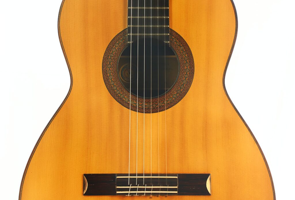 Nahaufnahme des Korpus einer „Manuel Adalid Sr. 1971 Francisco“ Klassischen Gitarre, die sechs Saiten, das Schallloch mit einer dekorativen Rosette und einen Steg aus Holz zeigt; fotografiert vor weißem Hintergrund. Nahaufnahme des Korpus einer „Manuel Adalid Sr. 1971 Francisco“ Klassischen Gitarre, die sechs Saiten, das Schallloch mit einer dekorativen Rosette und einen Steg aus Holz zeigt; fotografiert vor weißem Hintergrund.