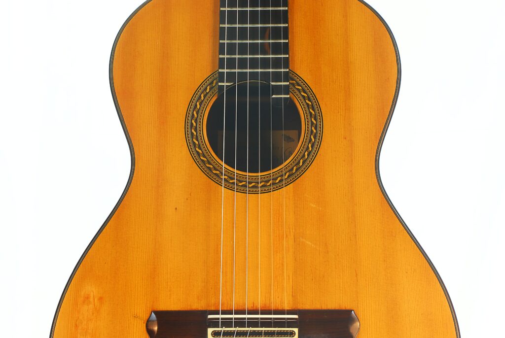 Eine Nahaufnahme des Korpus der „Antonius Müller 2004 Doubletop Klassische Gitarre“, die die Holzoberfläche, das kunstvoll verzierte Schallloch und die sechs Saiten vor weißem Hintergrund hervorhebt. Eine Nahaufnahme des Korpus der „Antonius Müller 2004 Doubletop Klassische Gitarre“, die die Holzoberfläche, das kunstvoll verzierte Schallloch und die sechs Saiten vor weißem Hintergrund hervorhebt.