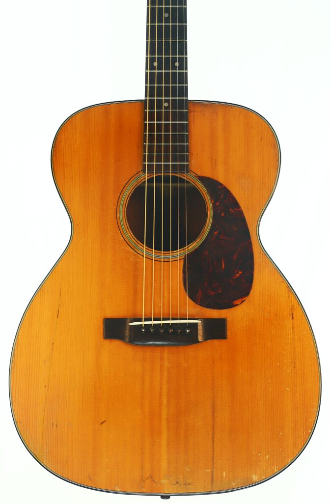 Nahaufnahme einer Martin 000-18 Akustikgitarre aus dem Jahr 1960 mit Holzkorpus, dunklem Schlagbrett und sechs Saiten, fotografiert vor weißem Hintergrund. Nahaufnahme einer Martin 000-18 Akustikgitarre aus dem Jahr 1960 mit Holzkorpus, dunklem Schlagbrett und sechs Saiten, fotografiert vor weißem Hintergrund.