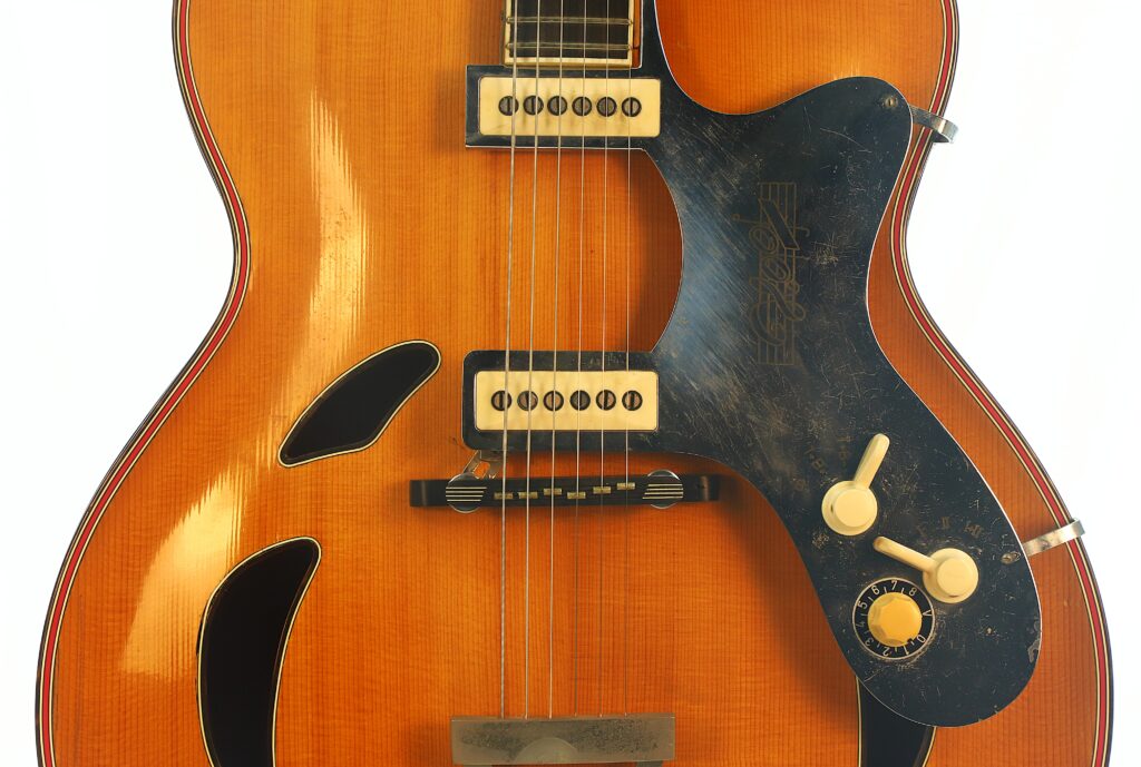 Nahaufnahme einer Artur-Lang-Jazzgitarre aus den 1950er Jahren, auf der zwei cremefarbene Tonabnehmer, ein schwarzes Schlagbrett mit sichtbaren Kratzern, Lautstärke- und Klangregler sowie f-förmige Schalllöcher auf einer glänzenden Holzoberfläche zu sehen sind. Nahaufnahme einer Artur-Lang-Jazzgitarre aus den 1950er Jahren, auf der zwei cremefarbene Tonabnehmer, ein schwarzes Schlagbrett mit sichtbaren Kratzern, Lautstärke- und Klangregler sowie f-förmige Schalllöcher auf einer glänzenden Holzoberfläche zu sehen sind.