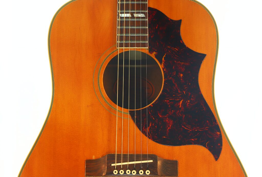 Nahaufnahme des Korpus einer Gibson SJN 1968 Akustikgitarre, bei der das Schallloch, die Saiten, der Holzsteg und ein großes, wellenförmiges, dunkles Schlagbrett vor dem Hintergrund der warmbraunen Lackierung besonders hervorgehoben werden. Nahaufnahme des Korpus einer Gibson SJN 1968 Akustikgitarre, bei der das Schallloch, die Saiten, der Holzsteg und ein großes, wellenförmiges, dunkles Schlagbrett vor dem Hintergrund der warmbraunen Lackierung besonders hervorgehoben werden.