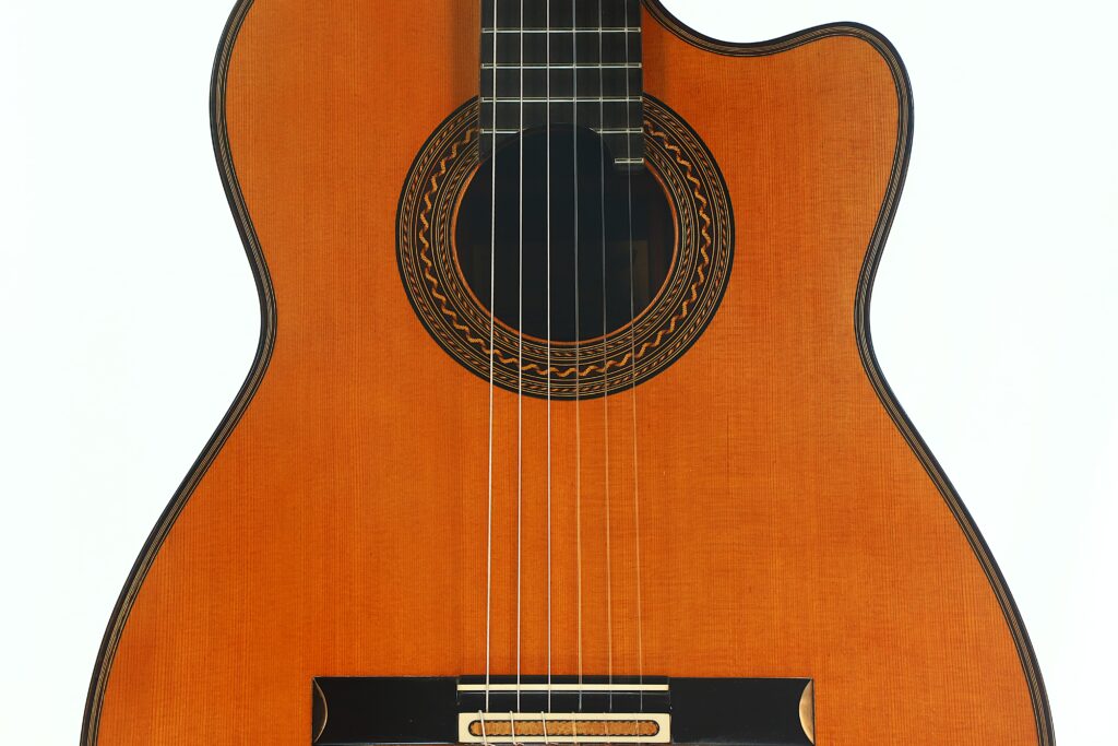 Nahaufnahme des Korpus der Antonius Müller 2012 Doubletop Klassische Gitarre, die die Holzoberfläche, die detailreiche Rosette um das Schallloch sowie die sechs sichtbaren Saiten vor weißem Hintergrund zeigt. Nahaufnahme des Korpus der Antonius Müller 2012 Doubletop Klassische Gitarre, die die Holzoberfläche, die detailreiche Rosette um das Schallloch sowie die sechs sichtbaren Saiten vor weißem Hintergrund zeigt.