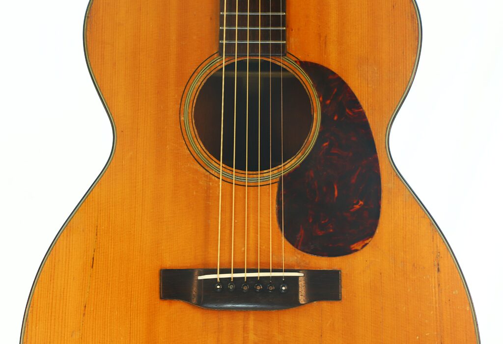 Nahaufnahme des Korpus der Martin 000-18 Akustikgitarre aus dem Jahr 1960, bei der das Schallloch, die Saiten, der Holzsteg und ein dunkles, tropfenförmiges Schlagbrett vor dem Hintergrund einer naturbelassenen Holzoberfläche besonders hervorgehoben werden. Nahaufnahme des Korpus der Martin 000-18 Akustikgitarre aus dem Jahr 1960, bei der das Schallloch, die Saiten, der Holzsteg und ein dunkles, tropfenförmiges Schlagbrett vor dem Hintergrund einer naturbelassenen Holzoberfläche besonders hervorgehoben werden.
