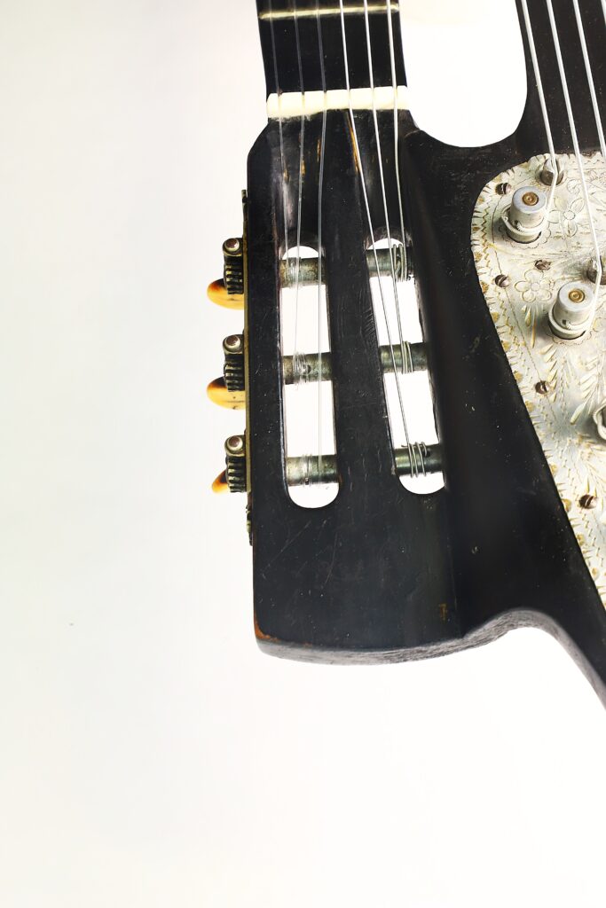 Nahaufnahme des Kopfstücks und der Stimmwirbel einer Gustav Keller Stauffer Stil 1900 Kontragitarre, die einen Teil des schwarzen Korpus mit dekorativen Metallelementen vor weißem Hintergrund zeigt. Nahaufnahme des Kopfstücks und der Stimmwirbel einer Gustav Keller Stauffer Stil 1900 Kontragitarre, die einen Teil des schwarzen Korpus mit dekorativen Metallelementen vor weißem Hintergrund zeigt.