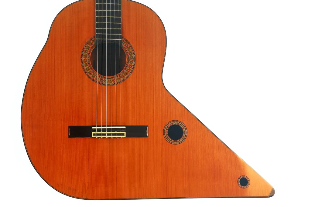 Die „Gran Klassische Gitarre“ von Raimundo y Aparicio Corujo ist eine Harfengitarre mit einem traditionellen Gitarrenhals und -korpus sowie einem verlängerten unteren Teil mit zwei zusätzlichen Schalllöchern; sie ist vor einem weißen Hintergrund abgebildet. Die „Gran Klassische Gitarre“ von Raimundo y Aparicio Corujo ist eine Harfengitarre mit einem traditionellen Gitarrenhals und -korpus sowie einem verlängerten unteren Teil mit zwei zusätzlichen Schalllöchern; sie ist vor einem weißen Hintergrund abgebildet.