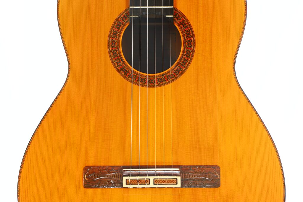 Nahaufnahme des Korpus einer Flamenco-Gitarre von Jeronimo Pena aus dem Jahr 1976, auf der sechs Saiten, ein geschnitzter Holzsteg und ein Rosettenmuster zu sehen sind, das das Schallloch umgibt, vor weißem Hintergrund. Nahaufnahme des Korpus einer Flamenco-Gitarre von Jeronimo Pena aus dem Jahr 1976, auf der sechs Saiten, ein geschnitzter Holzsteg und ein Rosettenmuster zu sehen sind, das das Schallloch umgibt, vor weißem Hintergrund.