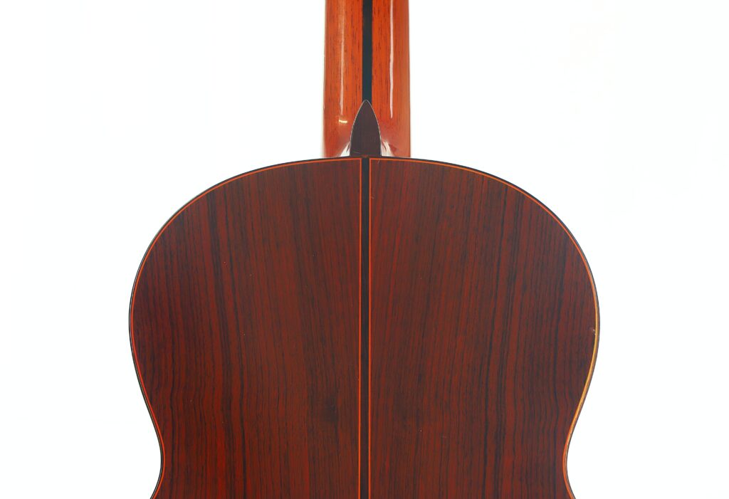 Nahaufnahme der Rückseite einer Jose Ramirez 1a 1977 Klassische Gitarre, die eine dunkle Holzmaserung mit einer vertikalen Mittelnaht sowie den sich vor weißem Hintergrund nach oben erstreckenden Hals zeigt. Nahaufnahme der Rückseite einer Jose Ramirez 1a 1977 Klassische Gitarre, die eine dunkle Holzmaserung mit einer vertikalen Mittelnaht sowie den sich vor weißem Hintergrund nach oben erstreckenden Hals zeigt.