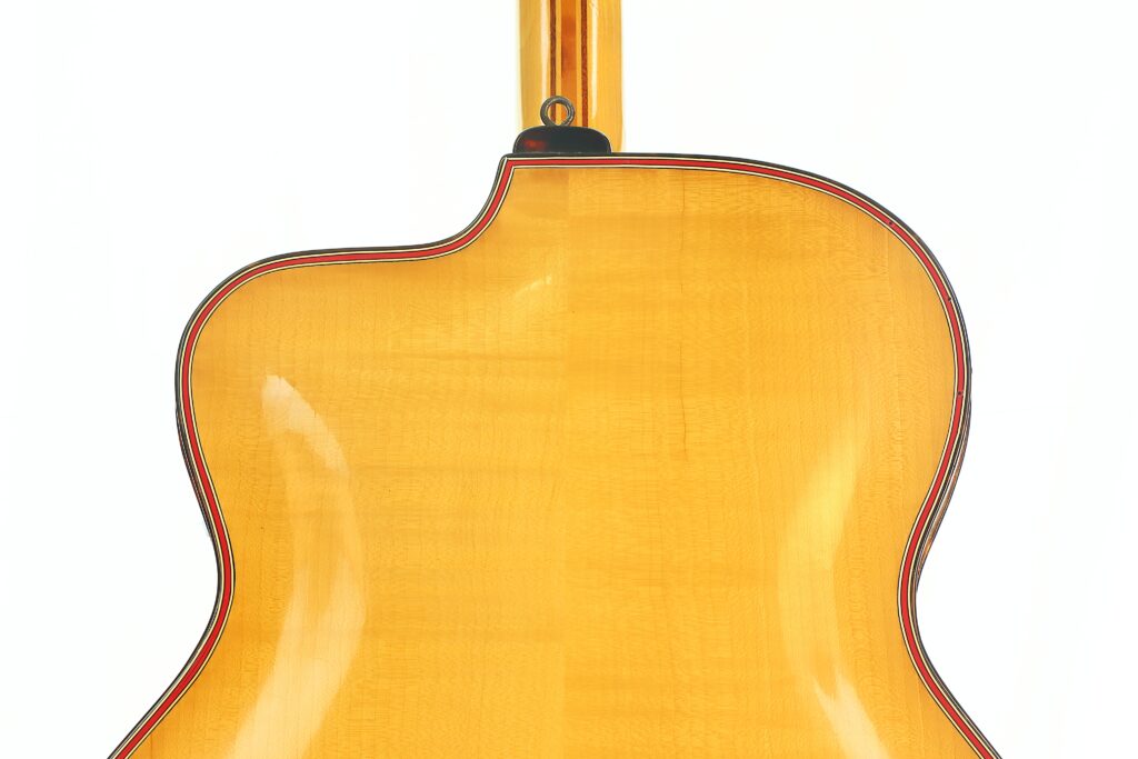 Nahaufnahme der Rückseite einer Artur-Lang-Jazzgitarre aus den 1950er Jahren mit einer glänzenden, hellen Holzoberfläche und einem einzelnen Cutaway nahe dem Hals. Die Gitarre ist an den Kanten mit einer schmalen schwarz-roten Umrandung verziert. Nahaufnahme der Rückseite einer Artur-Lang-Jazzgitarre aus den 1950er Jahren mit einer glänzenden, hellen Holzoberfläche und einem einzelnen Cutaway nahe dem Hals. Die Gitarre ist an den Kanten mit einer schmalen schwarz-roten Umrandung verziert.