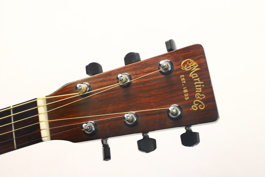 Nahaufnahme des Kopfes einer MARTIN OM-21 Akustikgitarre aus dem Jahr 1995, auf der sechs Stimmwirbel und das goldene „Martin & Co.“-Logo auf der rechten Seite zu sehen sind, vor einem schlichten weißen Hintergrund. Nahaufnahme des Kopfes einer MARTIN OM-21 Akustikgitarre aus dem Jahr 1995, auf der sechs Stimmwirbel und das goldene „Martin & Co.“-Logo auf der rechten Seite zu sehen sind, vor einem schlichten weißen Hintergrund.