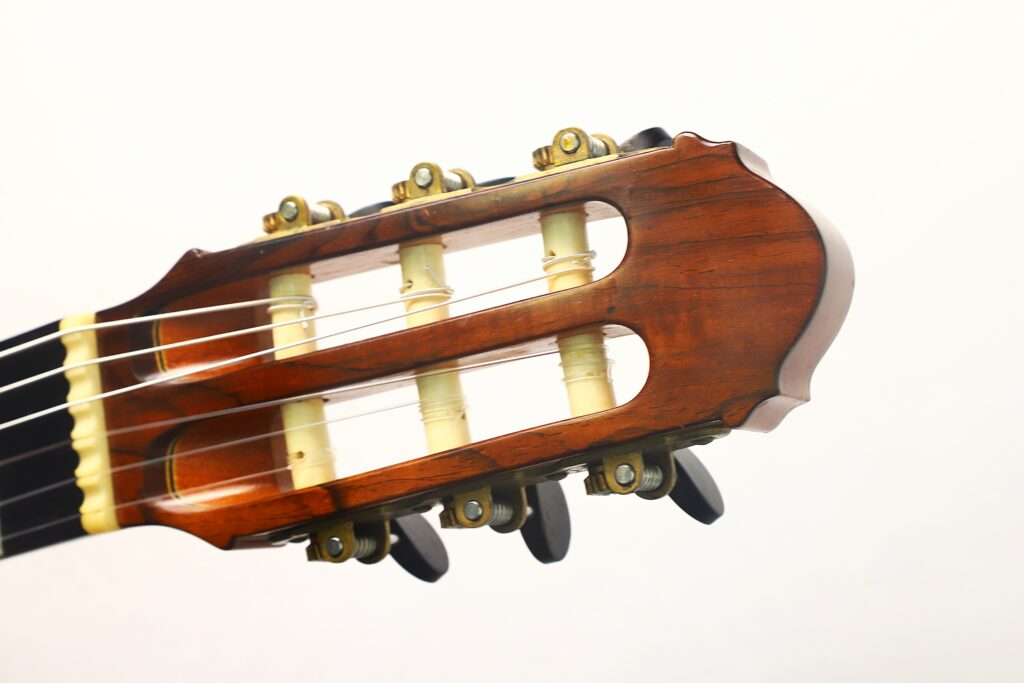 Nahaufnahme des Kopfstücks einer „Antonius Müller 2004 Doubletop Klassische Gitarre“ mit Stimmwirbeln und Nylonsaiten, wobei die Akzente aus Holz und elfenbeinähnlichem Material sich vom schlichten weißen Hintergrund abheben. Nahaufnahme des Kopfstücks einer „Antonius Müller 2004 Doubletop Klassische Gitarre“ mit Stimmwirbeln und Nylonsaiten, wobei die Akzente aus Holz und elfenbeinähnlichem Material sich vom schlichten weißen Hintergrund abheben.