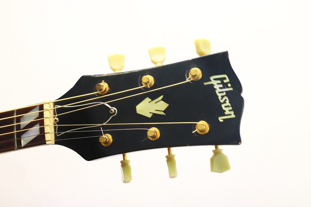 Nahaufnahme des Kopfstücks einer schwarzen Gibson SJN 1968 Akustikgitarre mit sechs Stimmwirbeln, goldfarbener Hardware und dem Gibson-Logo auf weißem Hintergrund. Nahaufnahme des Kopfstücks einer schwarzen Gibson SJN 1968 Akustikgitarre mit sechs Stimmwirbeln, goldfarbener Hardware und dem Gibson-Logo auf weißem Hintergrund.