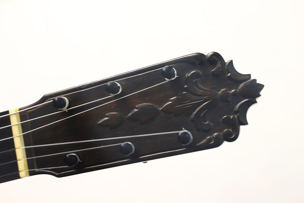 Nahaufnahme des Kopfstücks einer Flamenco-Gitarre von Jeronimo Pena aus dem Jahr 1976, gefertigt aus dunklem Holz und verziert mit einem kunstvollen, geschnitzten Blumenmuster sowie sechs Stimmwirbeln, vor weißem Hintergrund. Nahaufnahme des Kopfstücks einer Flamenco-Gitarre von Jeronimo Pena aus dem Jahr 1976, gefertigt aus dunklem Holz und verziert mit einem kunstvollen, geschnitzten Blumenmuster sowie sechs Stimmwirbeln, vor weißem Hintergrund.