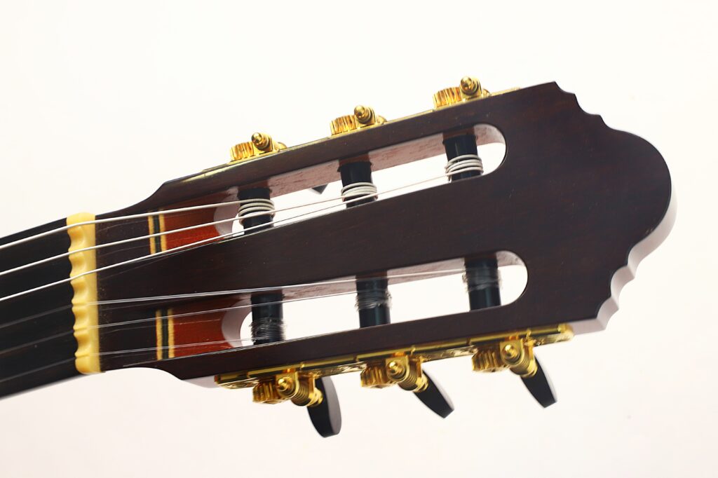 Nahaufnahme des Kopfes der „Antonius Müller 2012 Doubletop Klassische Gitarre“ mit goldenen Stimmwirbeln, weißen Saiten und einer dunklen Holzoberfläche, abgebildet vor einem schlichten weißen Hintergrund. Nahaufnahme des Kopfes der „Antonius Müller 2012 Doubletop Klassische Gitarre“ mit goldenen Stimmwirbeln, weißen Saiten und einer dunklen Holzoberfläche, abgebildet vor einem schlichten weißen Hintergrund.