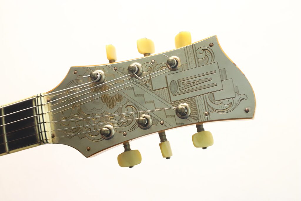 Nahaufnahme der Kopfplatte der Artur-Lang-Jazzgitarre aus den 1950er Jahren mit kunstvollen Metallgravuren, sechs Stimmwirbeln mit cremefarbenen Knöpfen und aufgespannten Saiten vor einem schlichten, hellen Hintergrund. Nahaufnahme der Kopfplatte der Artur-Lang-Jazzgitarre aus den 1950er Jahren mit kunstvollen Metallgravuren, sechs Stimmwirbeln mit cremefarbenen Knöpfen und aufgespannten Saiten vor einem schlichten, hellen Hintergrund.