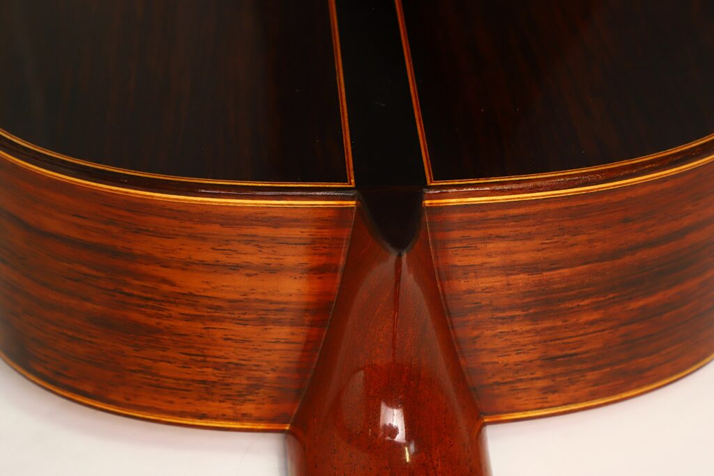 Nahaufnahme des Rücken- und Halsansatzes der „Friederich Klassische Gitarre“ von Stefan Nitschke aus dem Jahr 2015, die die detaillierte Maserung des Holzes, einen dunkelbraunen Boden, hellbraune Zargen und eine feine gelbe Einfassung zeigt. Nahaufnahme des Rücken- und Halsansatzes der „Friederich Klassische Gitarre“ von Stefan Nitschke aus dem Jahr 2015, die die detaillierte Maserung des Holzes, einen dunkelbraunen Boden, hellbraune Zargen und eine feine gelbe Einfassung zeigt.